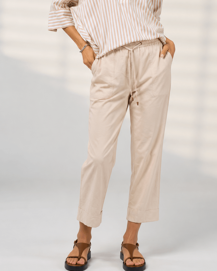 Madly Sweetly model  wearing light beige drawstring cropped pants with a striped shirt and brown sandals, standing against a light gray studio background with soft window-like shadows.