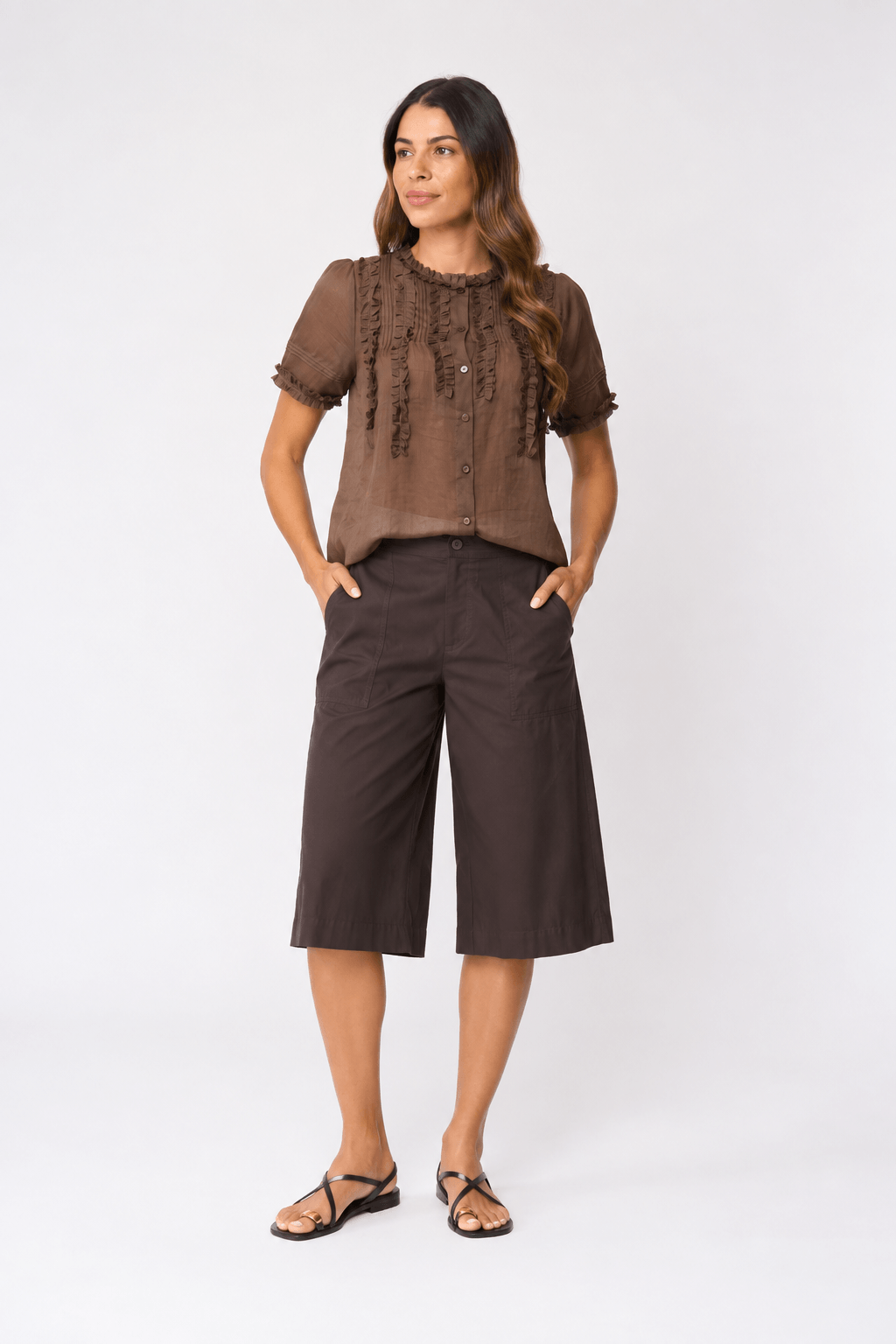 Foil clothing modeling a chocolate fril front and dark brown wide-leg cropped culottes with black strappy sandals, standing against a light gray studio background.