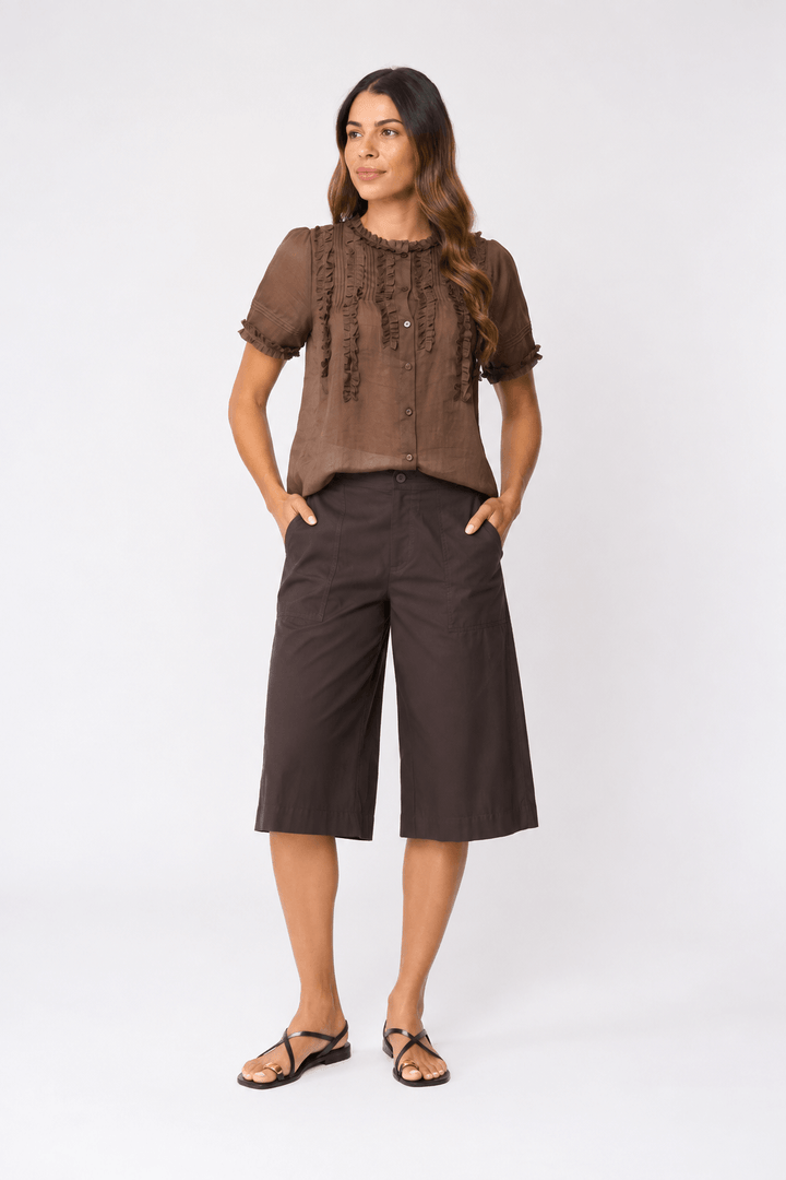 Foil clothing modeling a chocolate fril front and dark brown wide-leg cropped culottes with black strappy sandals, standing against a light gray studio background.