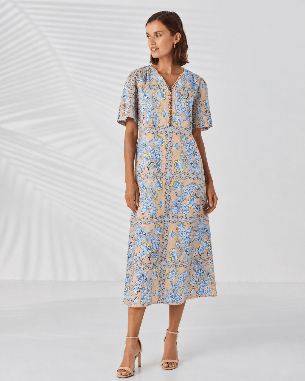 Lania The label model in a short-sleeve midi dress with a blue and beige paisley pattern, standing against a white studio background with leaf shadows.