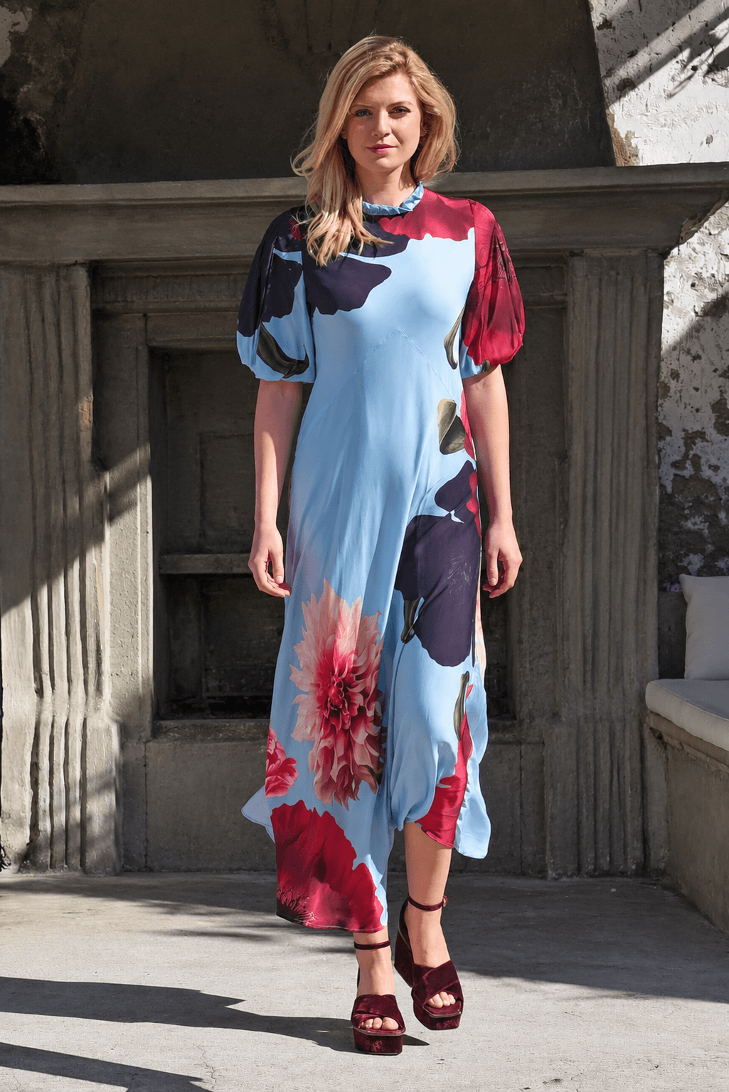Trelise Cooper models a light blue midi dress with a bold floral print in red, pink, and dark navy. The dress features short puff sleeves and a high neckline, paired with dark burgundy strappy platform sandals, set against a rustic stone background.