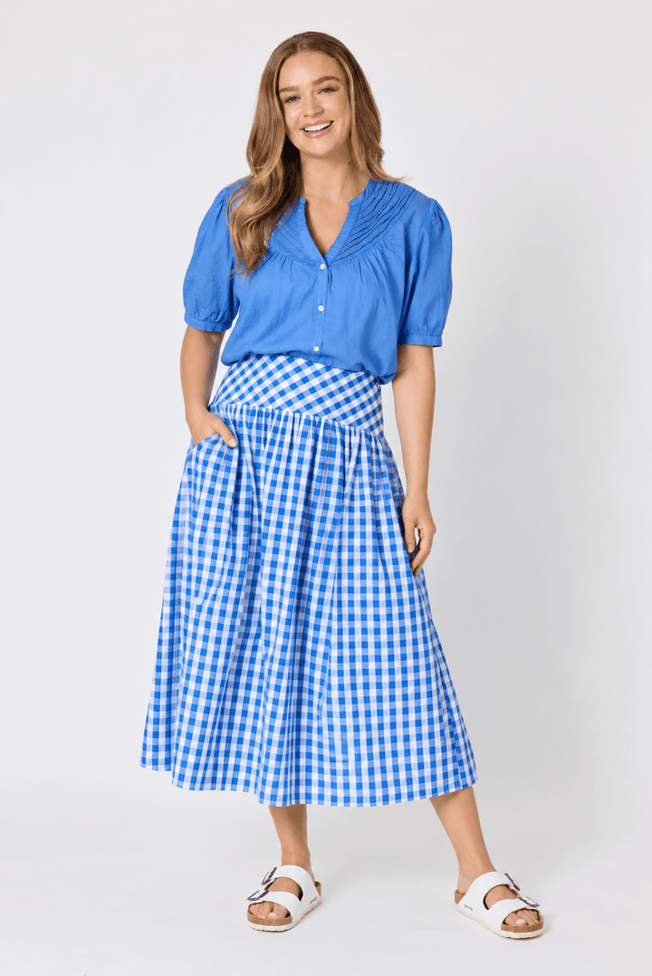 Threadz woman modeling a blue short-sleeve button-up blouse paired with a blue and white gingham midi skirt, styled with white slide sandals against a light gray studio background.