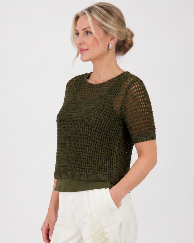 Monari  modeling a short-sleeve olive green knit top paired with white wide-leg pants featuring a green palm tree print, set against a plain light gray studio background.