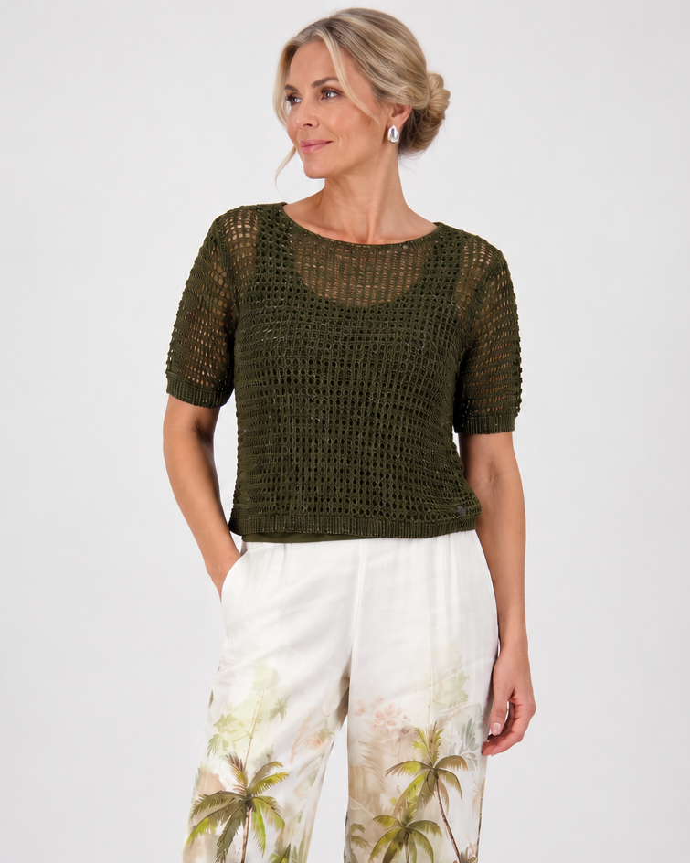 Monari  modeling a short-sleeve olive green knit top paired with white wide-leg pants featuring a green palm tree print, set against a plain light gray studio background.