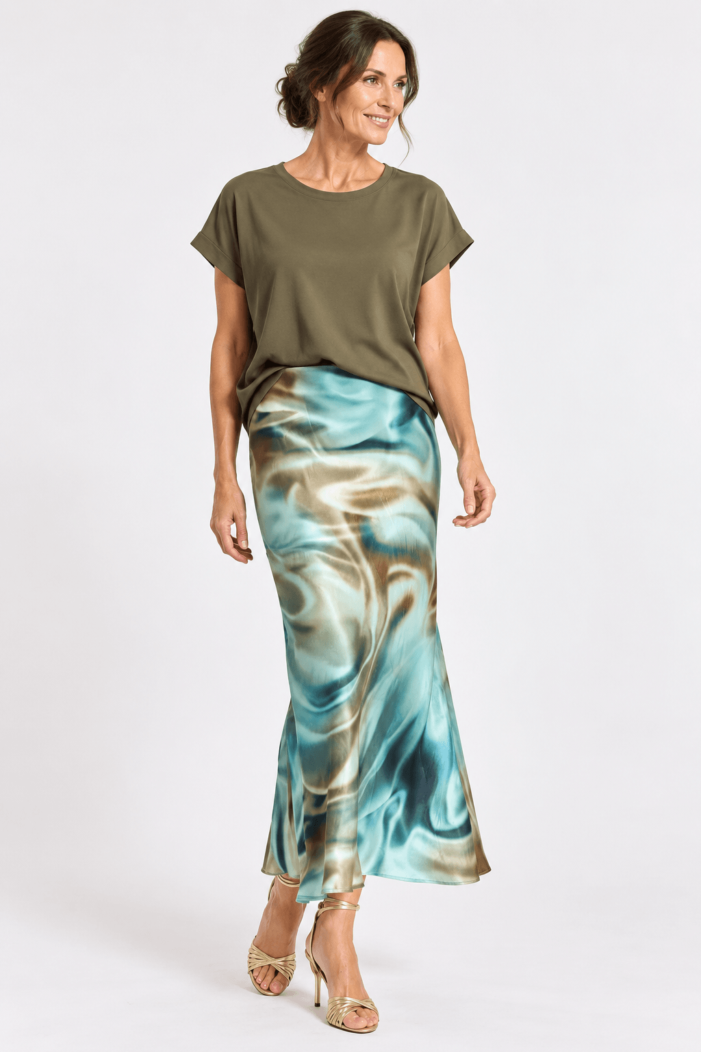 Lauren Vidal woman modeling a short-sleeve olive green top and a satin-like midi skirt with a blue, teal, and beige marbled pattern, paired with beige high heels, against a light gray studio background.