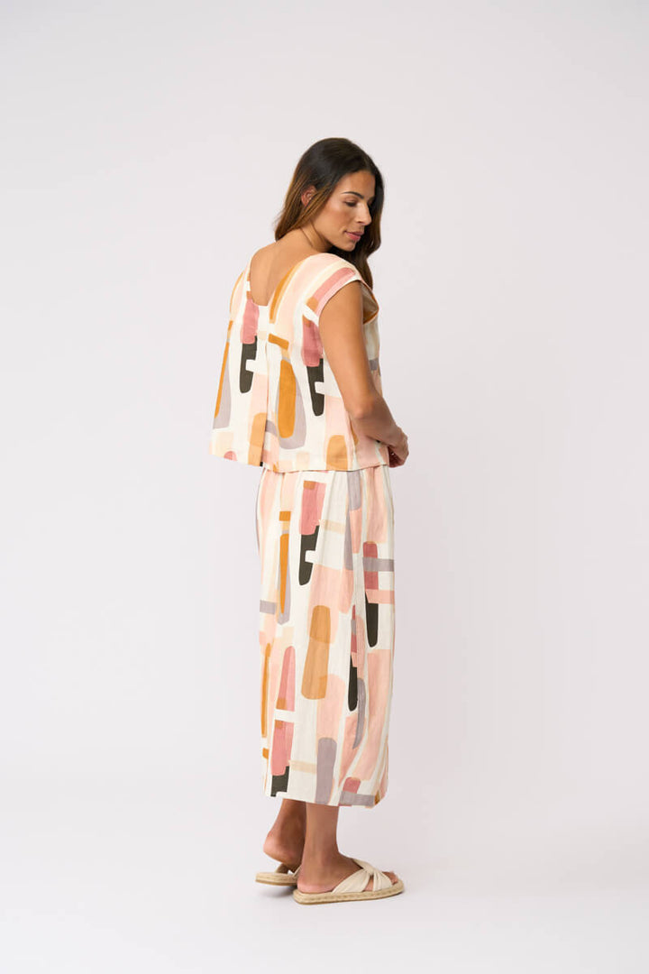 Foil modeling a sleeveless midi dress with a matching belt and skirt, featuring an abstract geometric print in peach, orange, pink, and beige on a white background, set against a plain light gray studio backdrop.
Back view 