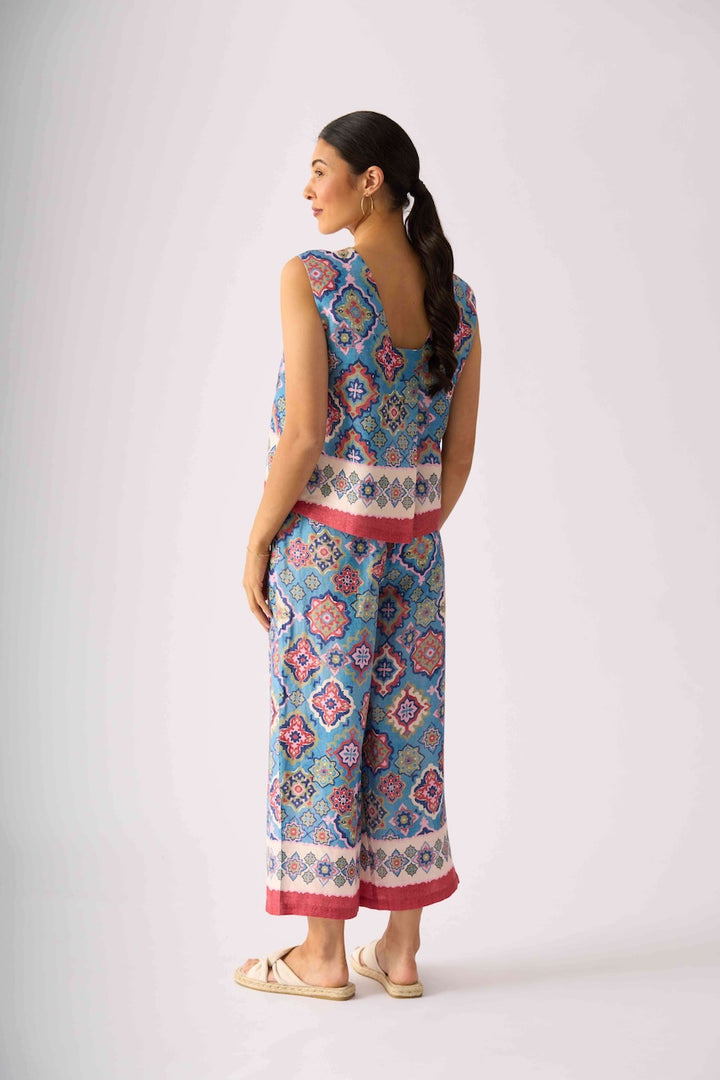 Foil modeling a sleeveless matching set of cropped tank top and wide-leg pants with a colorful geometric pattern in blue, pink, and white, paired with beige slide sandals, against a light gray studio background. Back view.