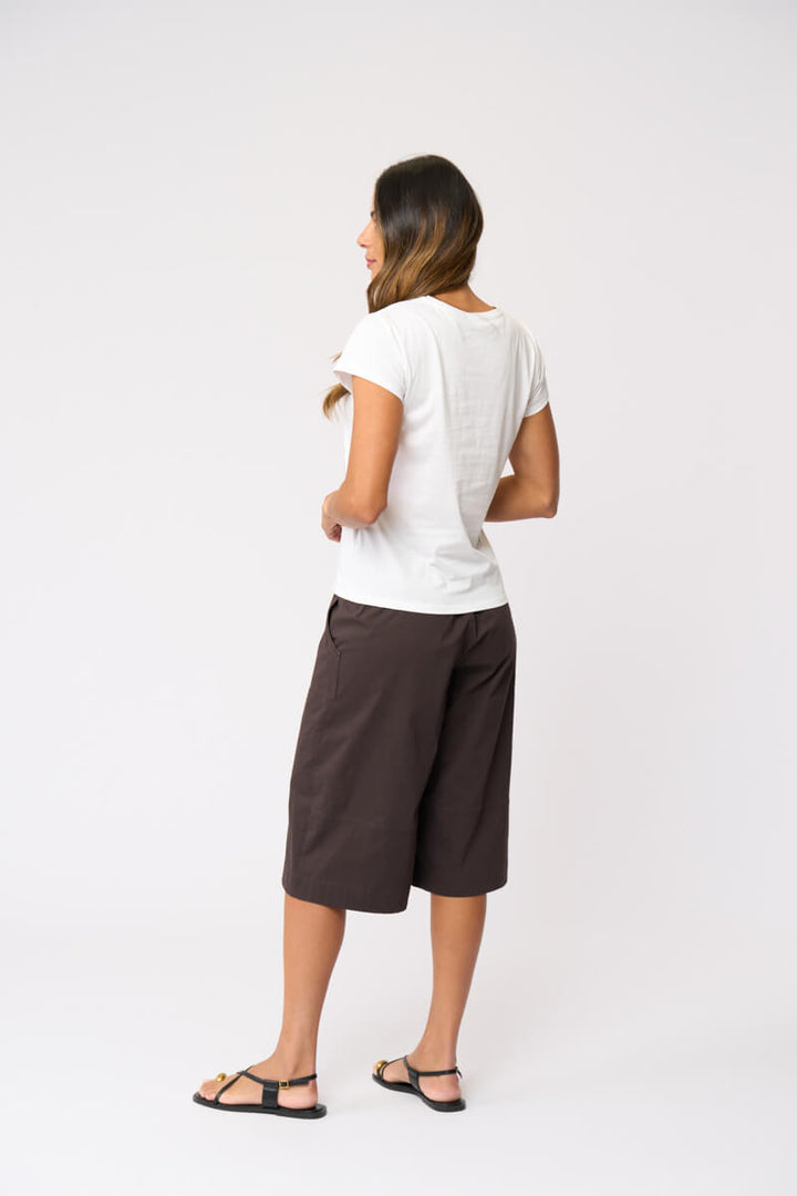 Foil clothing modeling a white short-sleeve t-shirt and dark brown wide-leg cropped pants with black strappy sandals, standing against a light gray studio background. Back view.