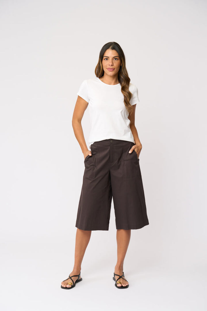 Foil clothing modeling a white short-sleeve t-shirt and dark brown wide-leg cropped pants with black strappy sandals, standing against a light gray studio background.