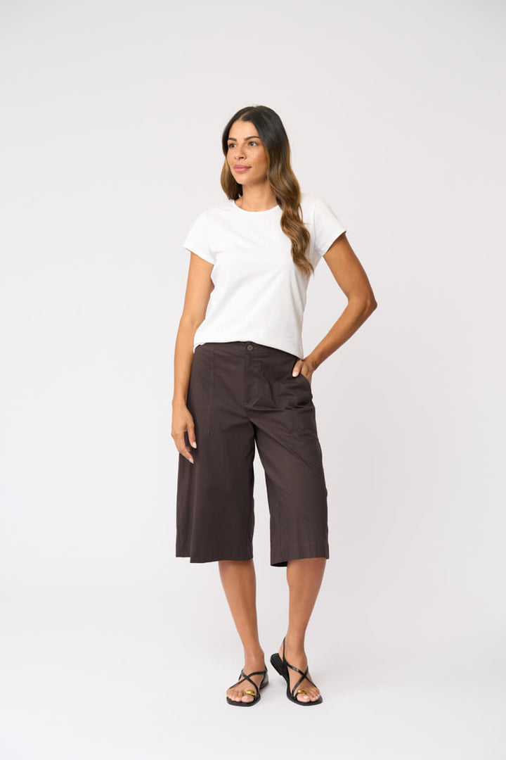 Foil clothing modeling a white short-sleeve t-shirt and dark brown wide-leg cropped pants with black strappy sandals, standing against a light gray studio background.