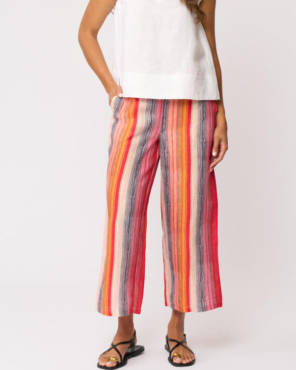 Foil model in a sleeveless white V-neck top and colorful vertically striped cropped pants in shades of red, orange, yellow, blue, and pink, paired with black strappy sandals, against a light gray studio background.