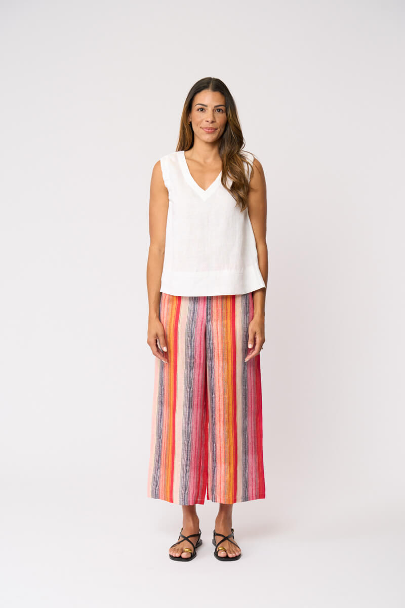 Foil model in a sleeveless white V-neck top and colorful vertically striped cropped pants in shades of red, orange, yellow, blue, and pink, paired with black strappy sandals, against a light gray studio background.