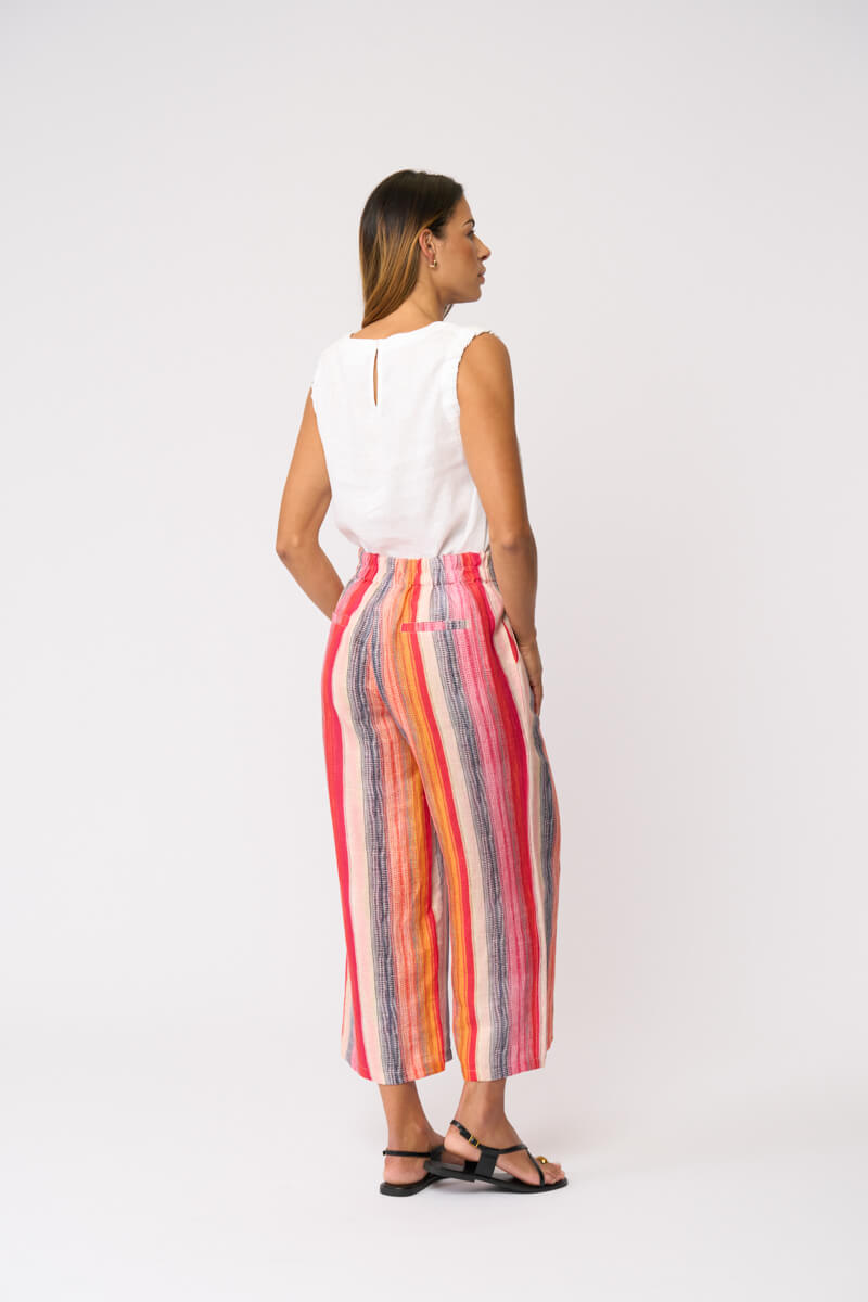 Foil model in a sleeveless white V-neck top and colorful vertically striped cropped pants in shades of red, orange, yellow, blue, and pink, paired with black strappy sandals, against a light gray studio background. Back view 