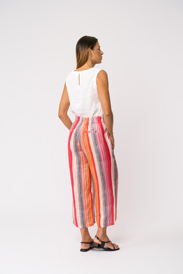Foil model in a sleeveless white V-neck top and colorful vertically striped cropped pants in shades of red, orange, yellow, blue, and pink, paired with black strappy sandals, against a light gray studio background. Back view 