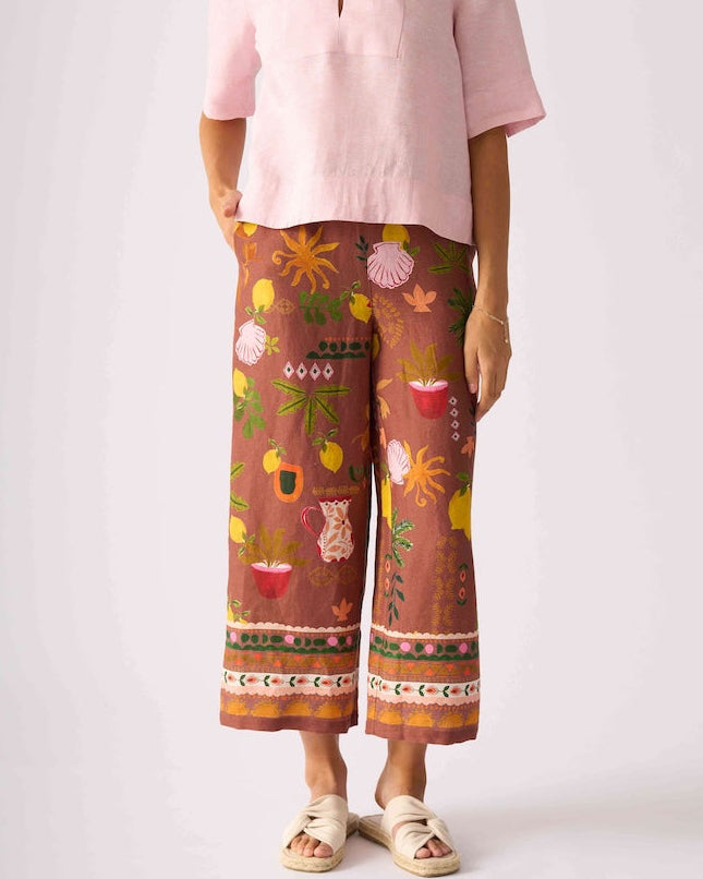 Foil clothing,  modeling a light pink short-sleeve top and brown wide-leg pants with a colorful floral and botanical print, standing against a light gray studio background.