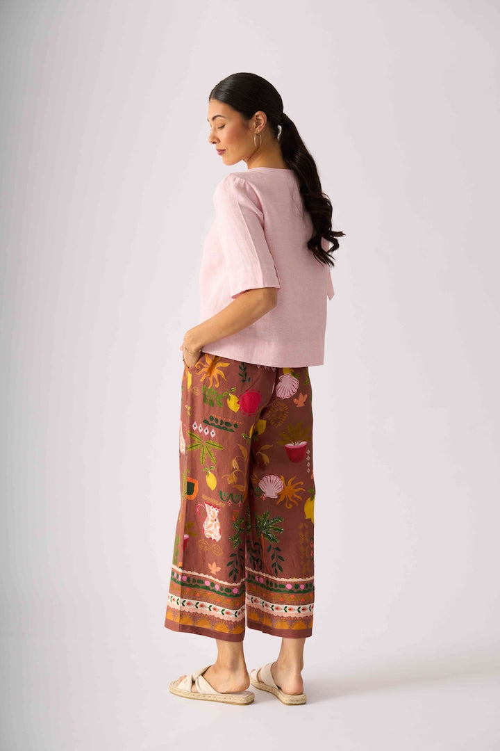 Foil clothing,  modeling a light pink short-sleeve top and brown wide-leg pants with a colorful floral and botanical print, standing against a light gray studio background.