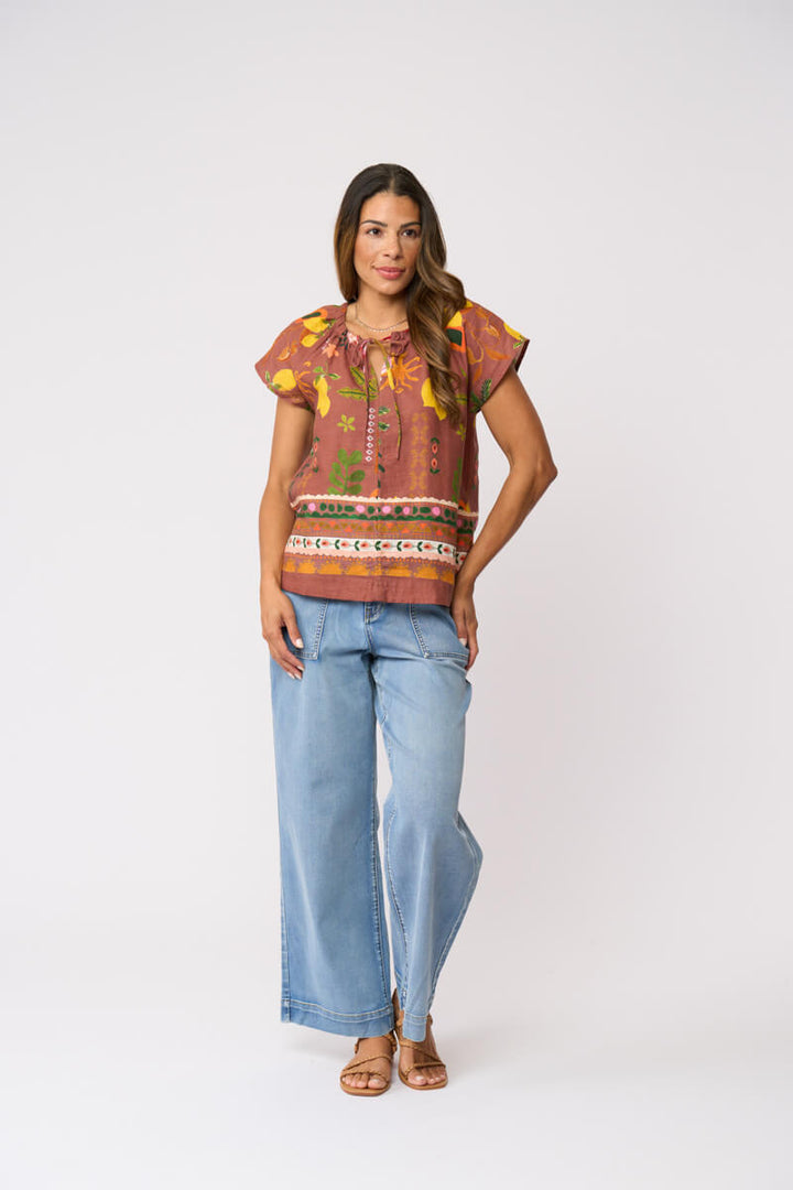 Foil Clothing, modeling a short-sleeve top with a brown, orange, and yellow patterned print, paired with light blue wide-leg jeans and brown sandals, against a plain light gray background.