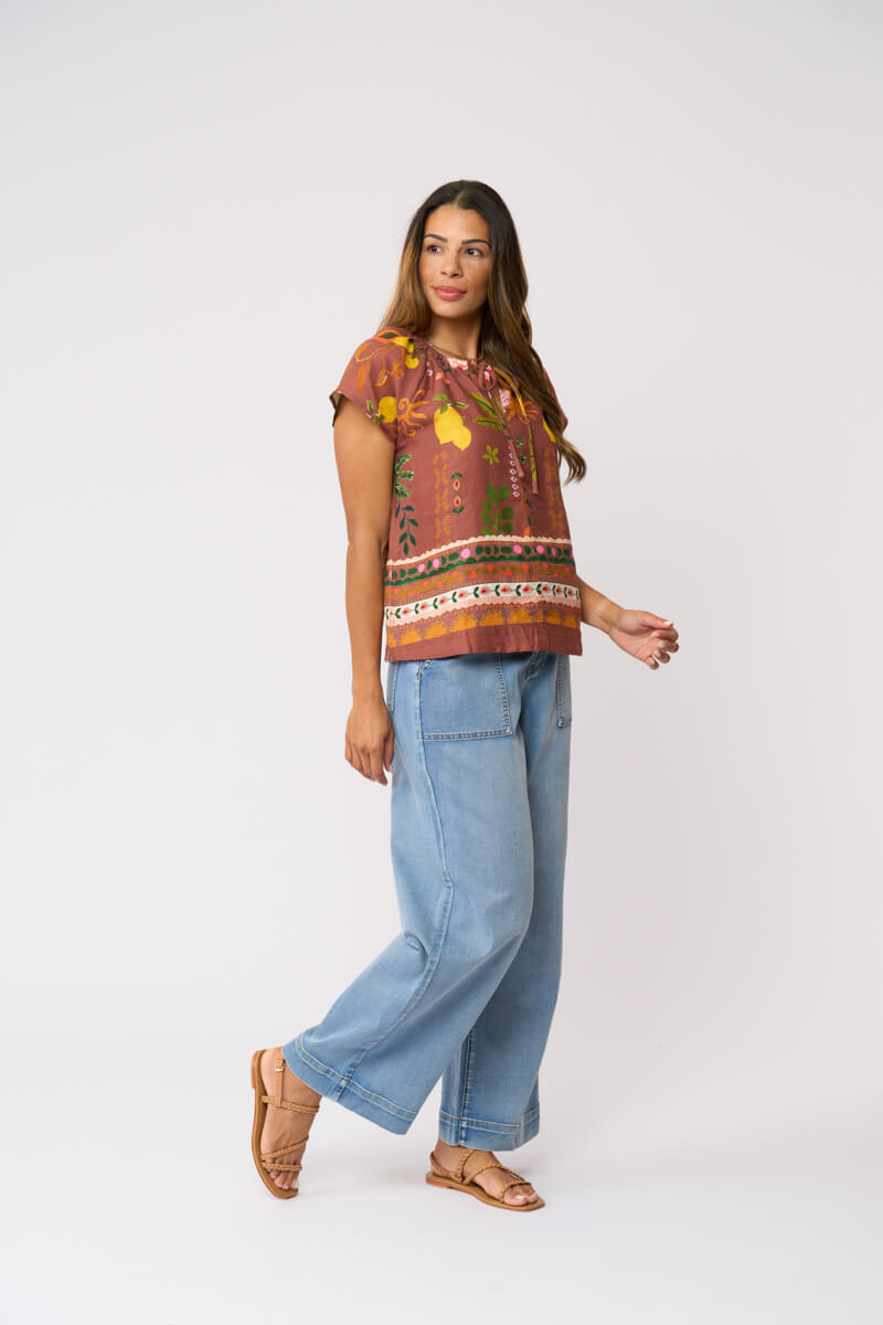 Foil Clothing, modeling a short-sleeve top with a brown, orange, and yellow patterned print, paired with light blue wide-leg jeans and brown sandals, against a plain light gray background.