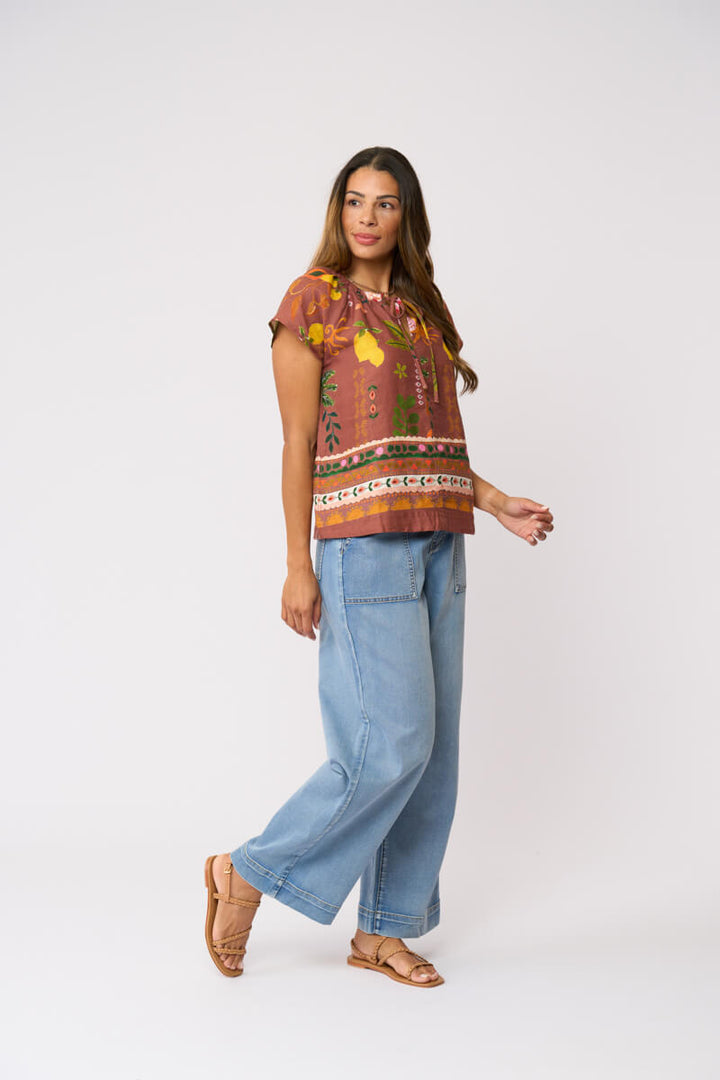 Foil Clothing, modeling a short-sleeve top with a brown, orange, and yellow patterned print, paired with light blue wide-leg jeans and brown sandals, against a plain light gray background.