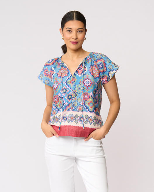 Foil modeling a short-sleeve patterned blouse with white cropped pants and tan sandals, standing against a plain light gray studio background.