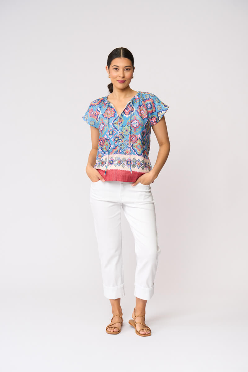Foil modeling a short-sleeve patterned blouse with white cropped pants and tan sandals, standing against a plain light gray studio background.