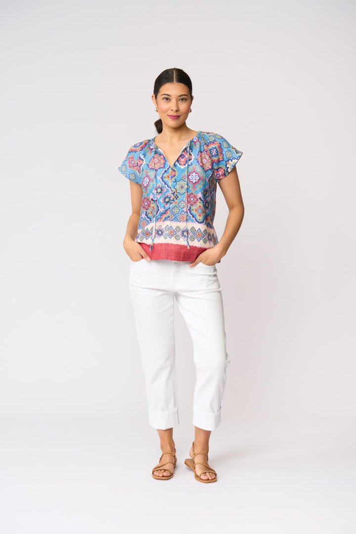 Foil modeling a short-sleeve patterned blouse with white cropped pants and tan sandals, standing against a plain light gray studio background.
