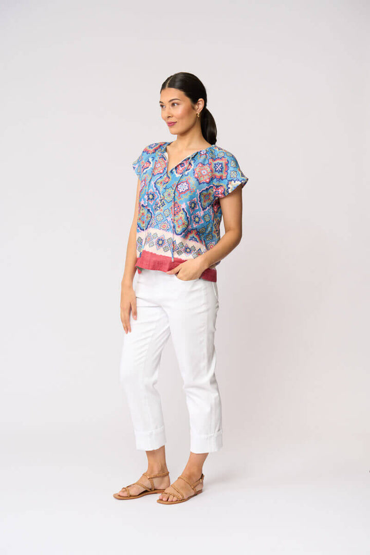 Foil modeling a short-sleeve patterned blouse with white cropped pants and tan sandals, standing against a plain light gray studio background.