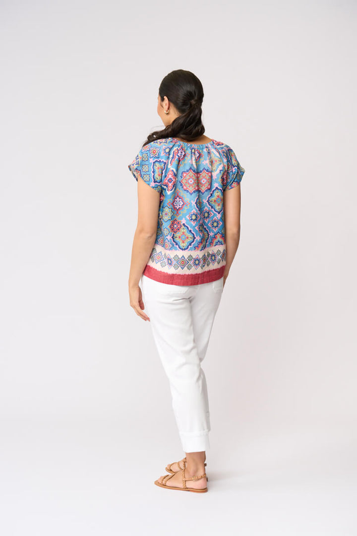 Foil modeling a short-sleeve patterned blouse with white cropped pants and tan sandals, standing against a plain light gray studio background. Back view.