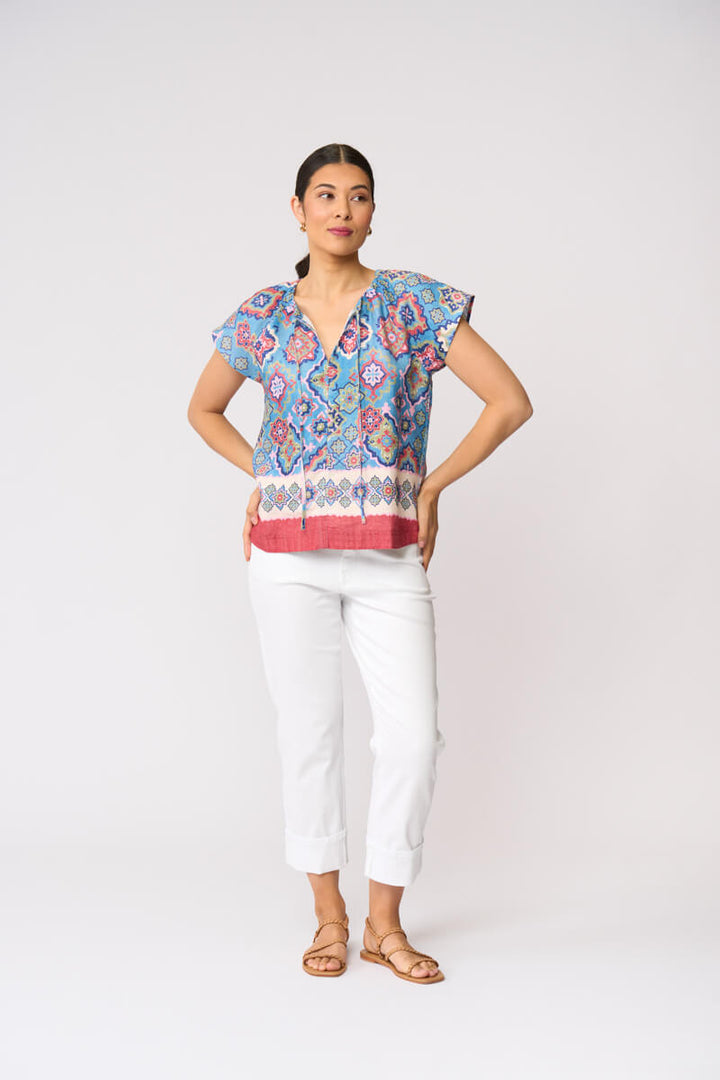 Foil modeling a short-sleeve patterned blouse with white cropped pants and tan sandals, standing against a plain light gray studio background.