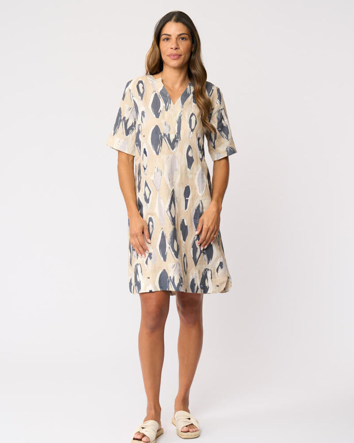 Foil modeling a short-sleeve V-neck dress with an abstract ikat pattern in beige and slate blue, paired with beige slide sandals, against a plain white studio background.