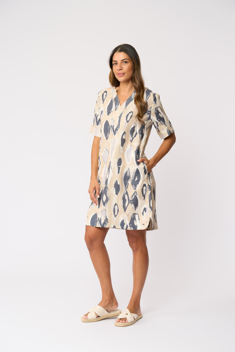 Foil modeling a short-sleeve V-neck dress with an abstract ikat pattern in beige and slate blue, paired with beige slide sandals, against a plain white studio background.