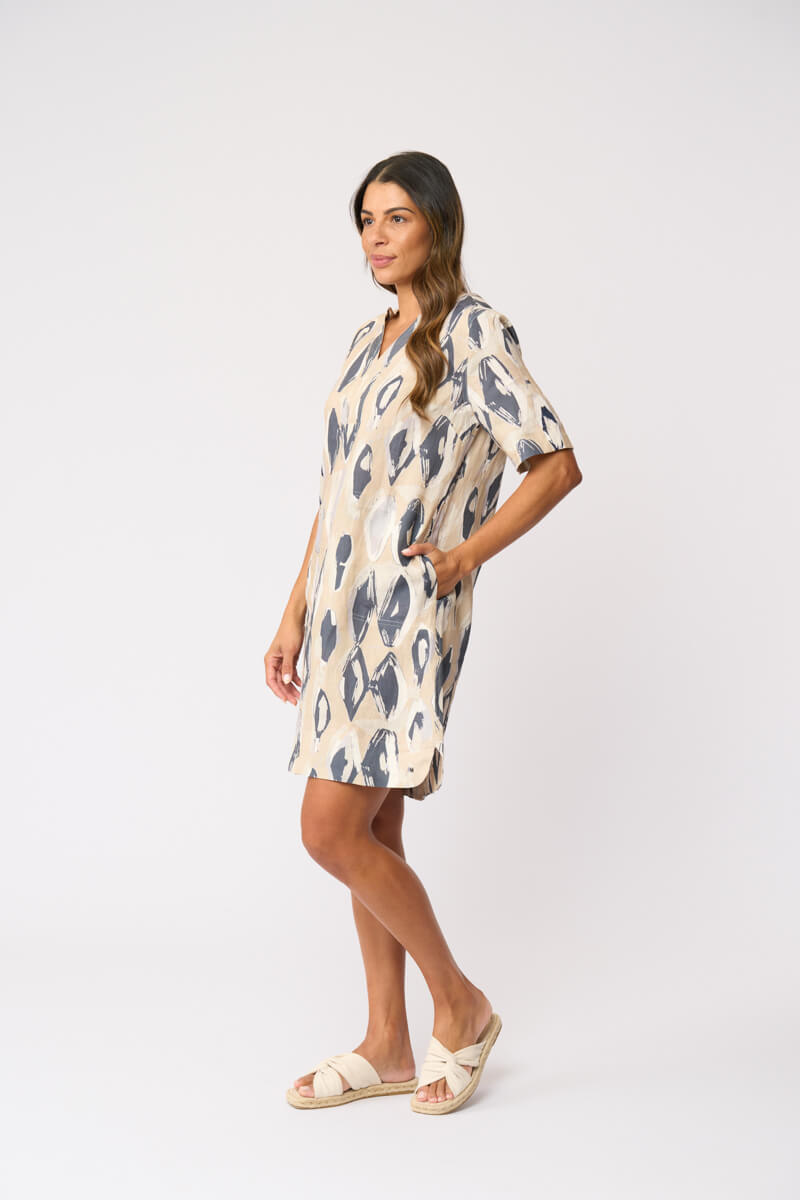 Foil modeling a short-sleeve V-neck dress with an abstract ikat pattern in beige and slate blue, paired with beige slide sandals, against a plain white studio background.