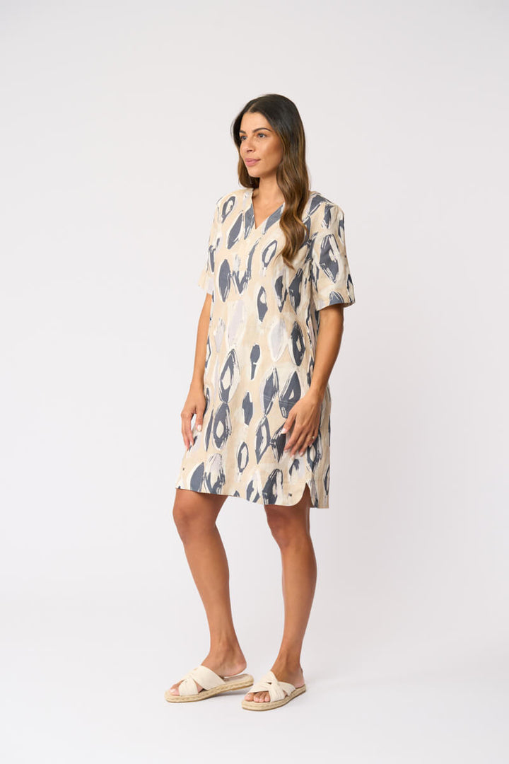 Foil modeling a short-sleeve V-neck dress with an abstract ikat pattern in beige and slate blue, paired with beige slide sandals, against a plain white studio background.