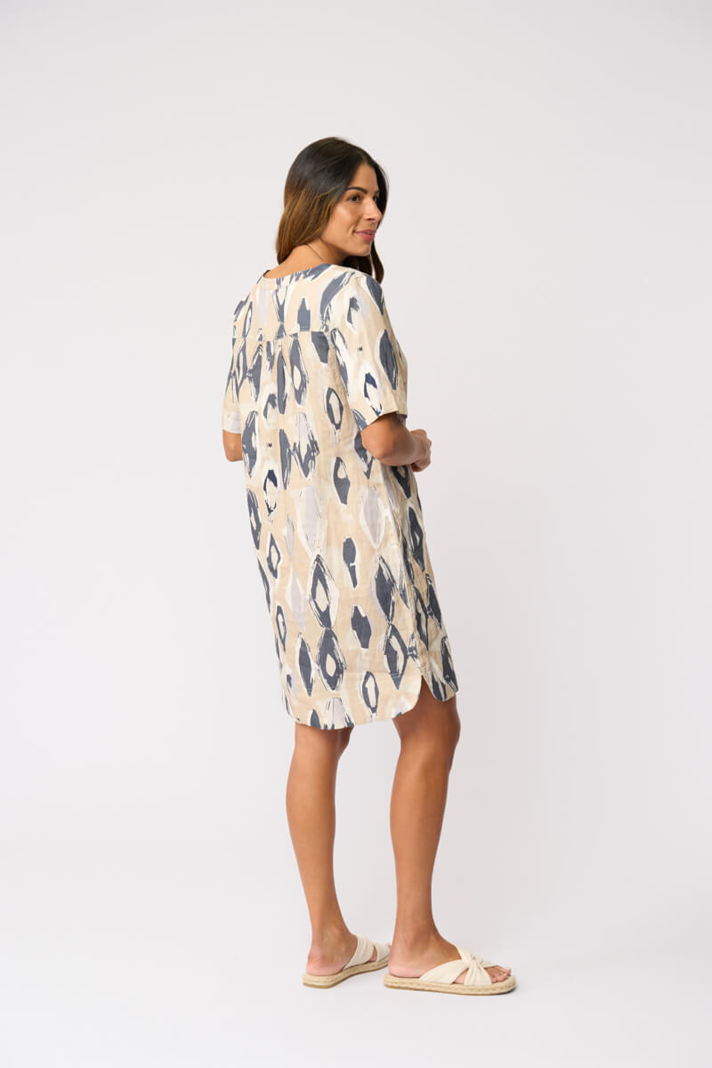 Foil modeling a short-sleeve V-neck dress with an abstract ikat pattern in beige and slate blue, paired with beige slide sandals, against a plain white studio background. Back view.