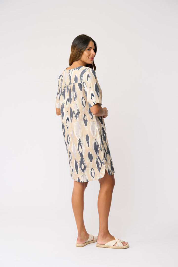 Foil modeling a short-sleeve V-neck dress with an abstract ikat pattern in beige and slate blue, paired with beige slide sandals, against a plain white studio background. Back view.