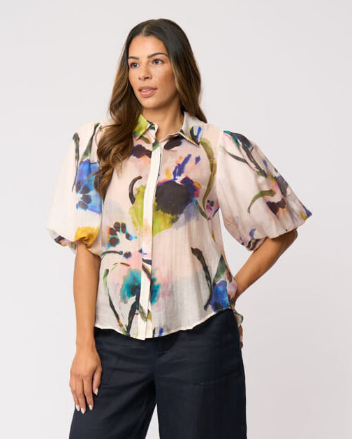 Foil clothing modeling a short-sleeve button-up blouse with a colorful abstract floral print on a light background, paired with dark cropped pants and beige heels, against a plain light gray studio background.