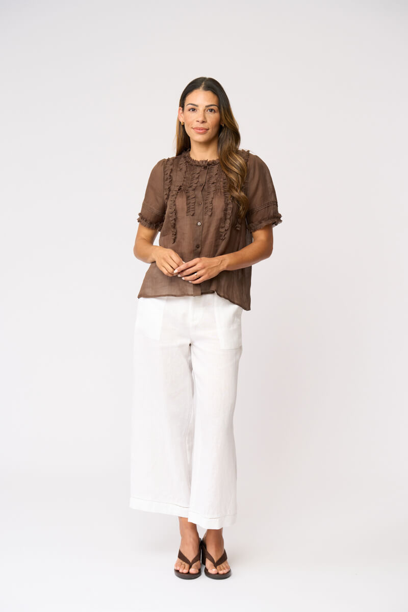 Foil clothing modeling a brown short-sleeve blouse with intricate lace and ruffle details on the front, paired with white wide-leg pants and black sandals, against a plain white studio backgroun