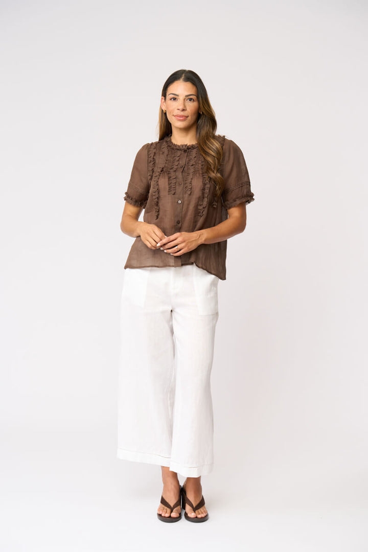 Foil clothing modeling a brown short-sleeve blouse with intricate lace and ruffle details on the front, paired with white wide-leg pants and black sandals, against a plain white studio backgroun