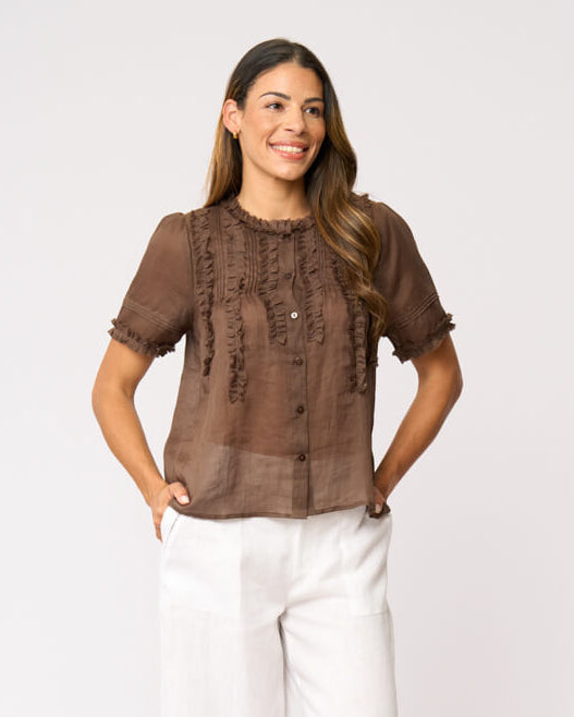 Foil clothing modeling a brown short-sleeve blouse with intricate lace and ruffle details on the front, paired with white wide-leg pants and black sandals, against a plain white studio background.