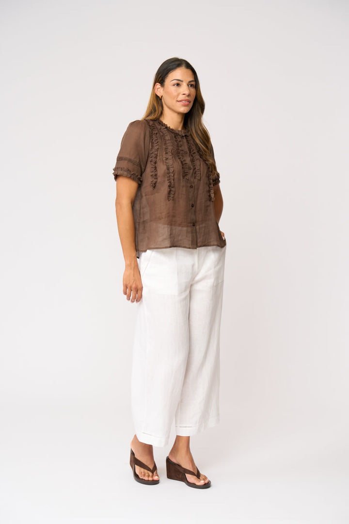 Foil clothing modeling a brown short-sleeve blouse with intricate lace and ruffle details on the front, paired with white wide-leg pants and black sandals, against a plain white studio backgroun