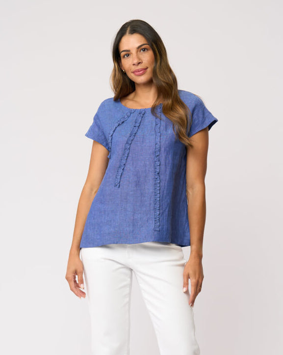 Foil clothing modeling a short-sleeve blue textured top with vertical stitched details, paired with white cropped pants and beige slide sandals, against a plain light gray studio background.