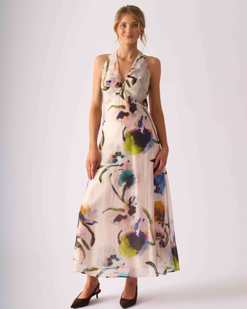 Foil Clothing model in  a sleeveless, wrap-style maxi dress with a multicolor floral watercolor print on a light cream background, paired with dark brown pointed-toe heels, against a plain light gray studio backdrop.