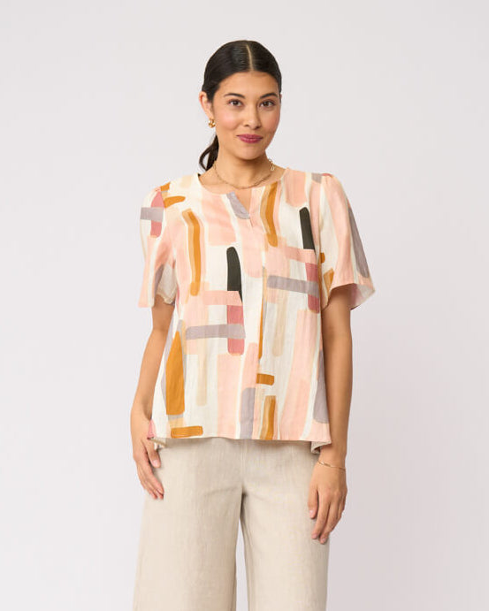 Foil model in a short-sleeve top with an abstract geometric print in peach, orange, beige, and gray, paired with wide-leg beige trousers and black strappy sandals, against a light gray studio background.