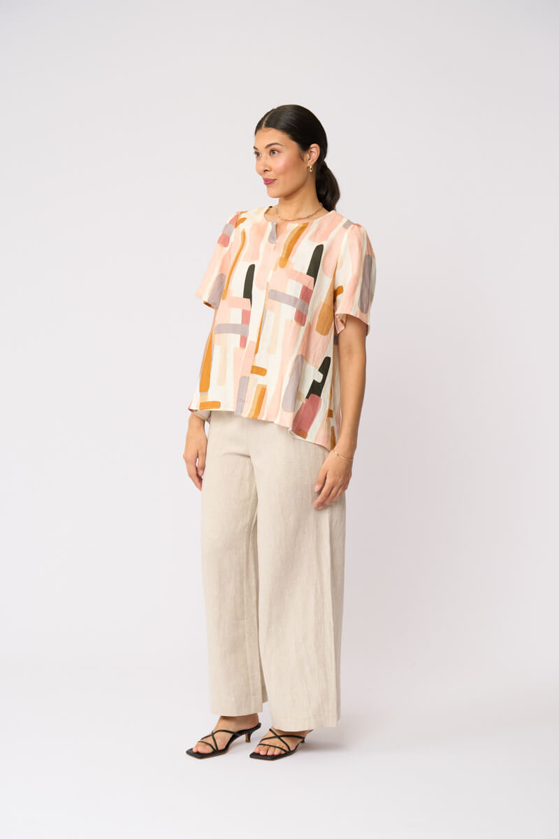 Foil model in a short-sleeve top with an abstract geometric print in peach, orange, beige, and gray, paired with wide-leg beige trousers and black strappy sandals, against a light gray studio background.