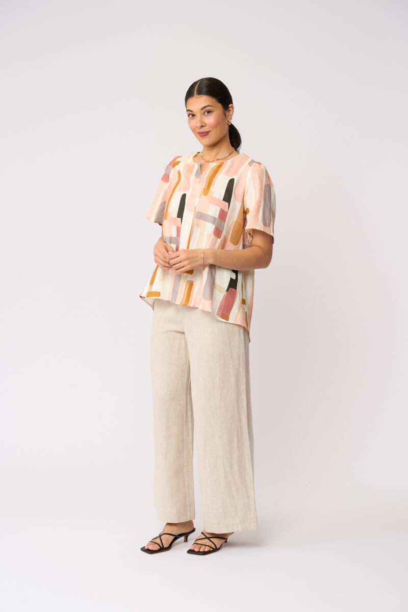 Foil model in a short-sleeve top with an abstract geometric print in peach, orange, beige, and gray, paired with wide-leg beige trousers and black strappy sandals, against a light gray studio background.