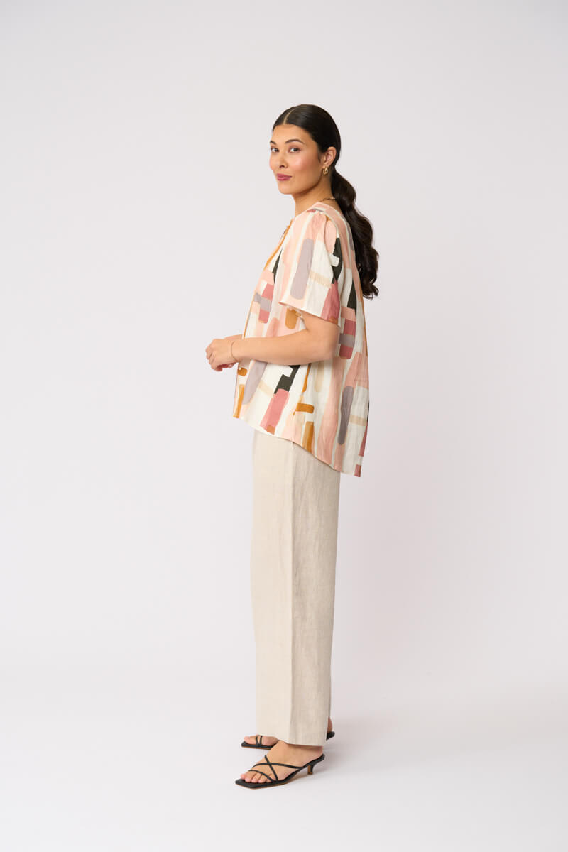 Foil model in a short-sleeve top with an abstract geometric print in peach, orange, beige, and gray, paired with wide-leg beige trousers and black strappy sandals, against a light gray studio background.
Side view