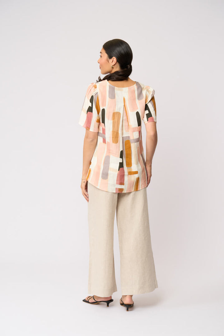 Foil model in a short-sleeve top with an abstract geometric print in peach, orange, beige, and gray, paired with wide-leg beige trousers and black strappy sandals, against a light gray studio background.
Back view