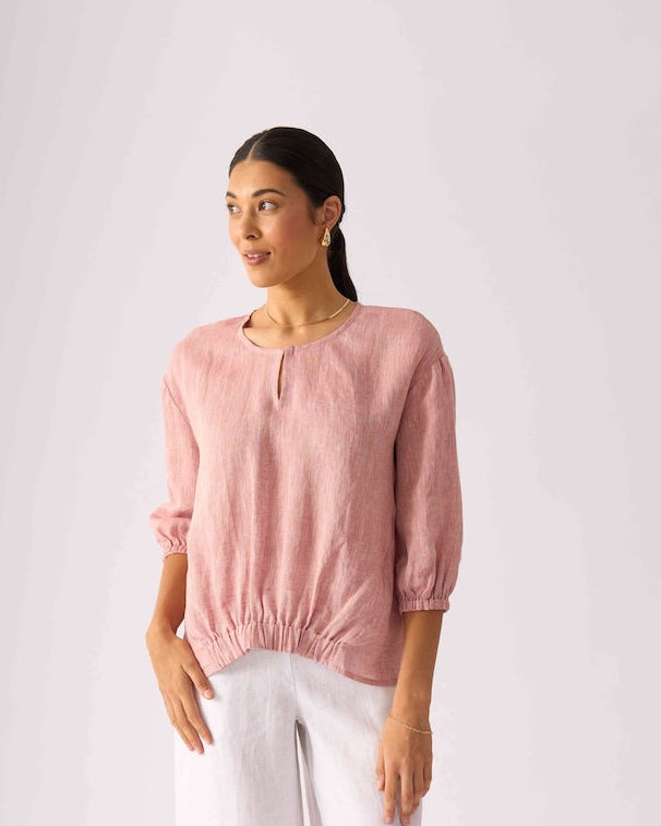 Foil Clothing modeling a dusty pink linen-blend top with puff sleeves and a gathered hem, paired with white wide-leg pants and beige sandals, against a light gray studio background.