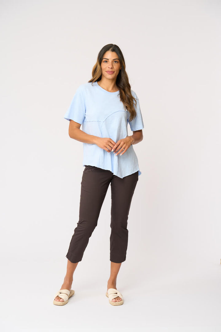 Foil clothing modeling a light blue short-sleeve top with asymmetrical seam details, paired with dark brown cropped pants and beige slide sandals, against a plain white studio background.