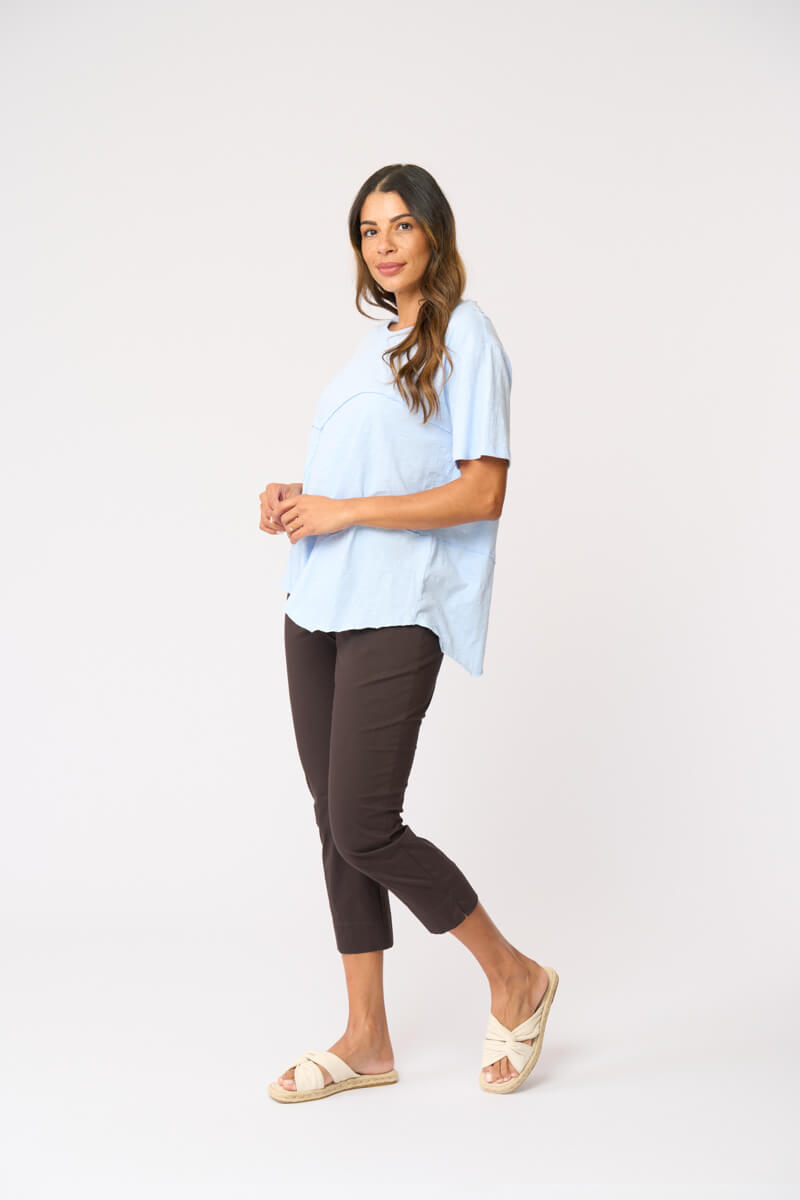 Foil clothing modeling a light blue short-sleeve top with asymmetrical seam details, paired with dark brown cropped pants and beige slide sandals, against a plain white studio background.